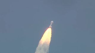 a rocket rises into a blue sky atop a conical pillar of flame.