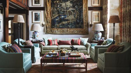 living room of a traditional jacobean home with wooden beams and a large tapestry hung on the wall, styled with artwork, a light green sofa, four tub chairs and a striped upholstered ottoman