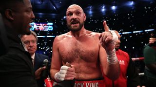 Tyson Fury, wearing red shorts emblazoned with 'Gypsy King', stands in a boxing ring pointing ahead of the Fury vs Usyk live stream 