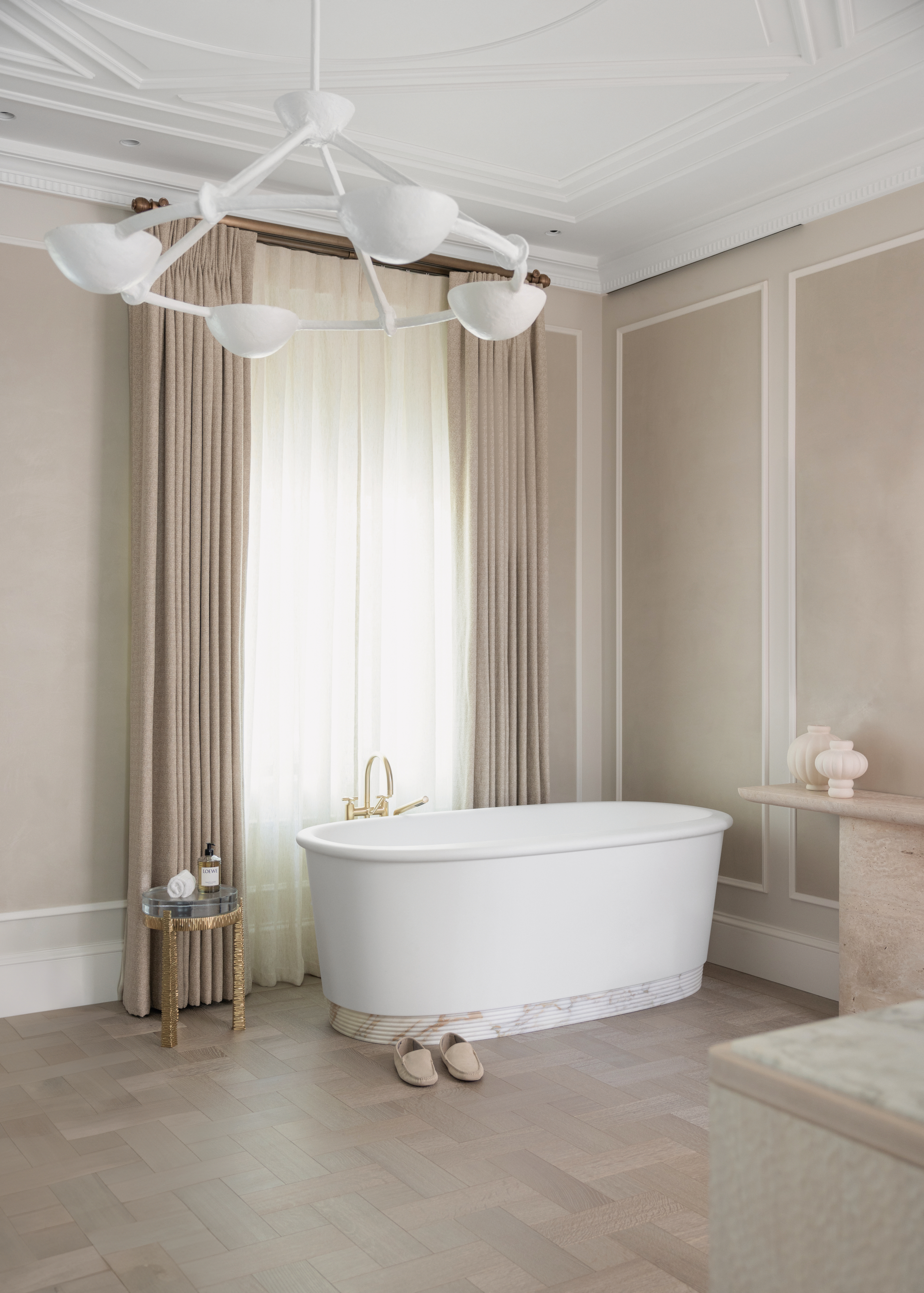 A freestanding white bathtub in the main bedroom with beige walls and beige curtains