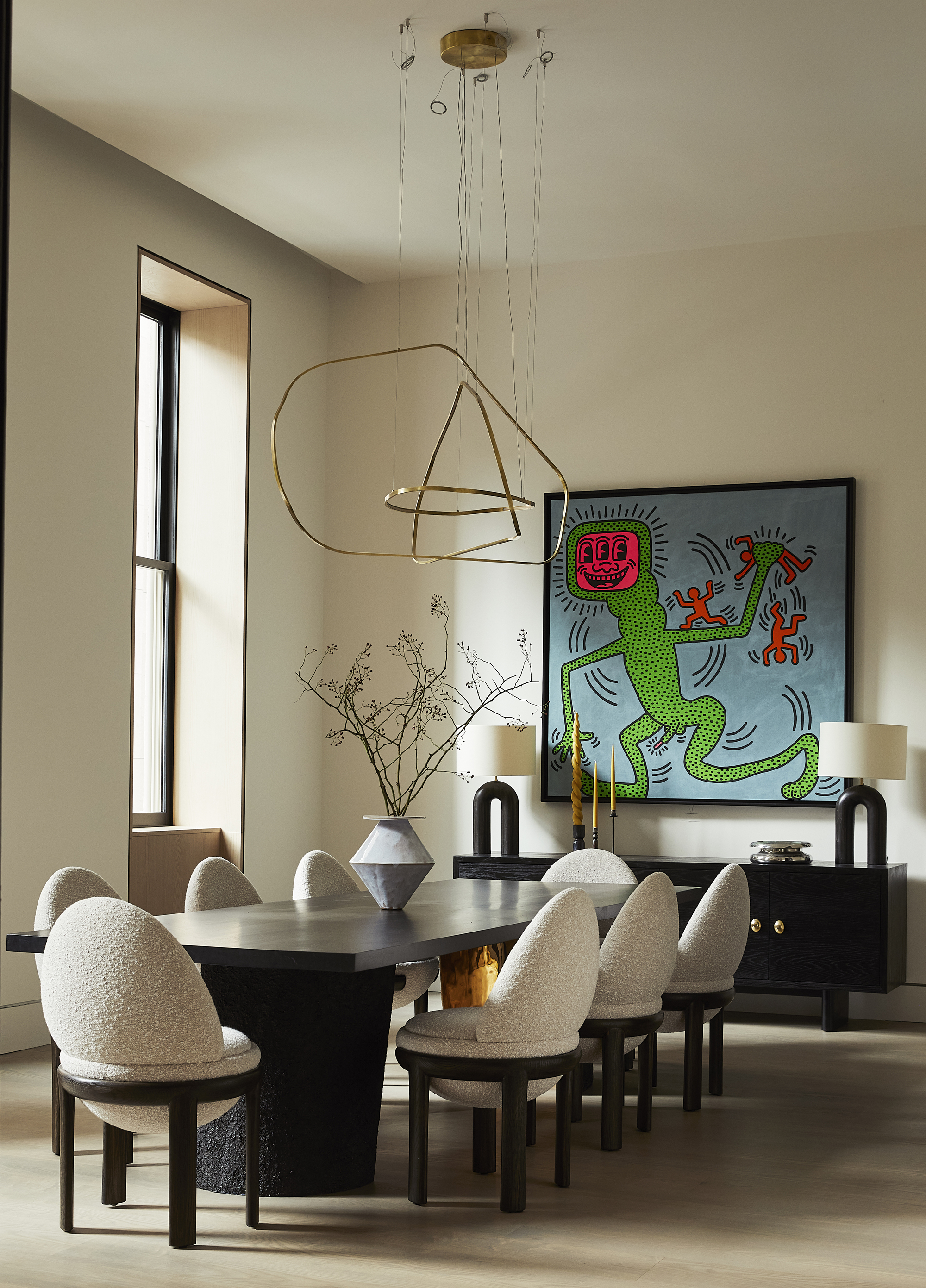 neutral dining room with long black dining table, boucle and black dining chairs, a vase on the table, a sculptural pendant light, a bright blue and green artwork hung over a black console at the end of the room