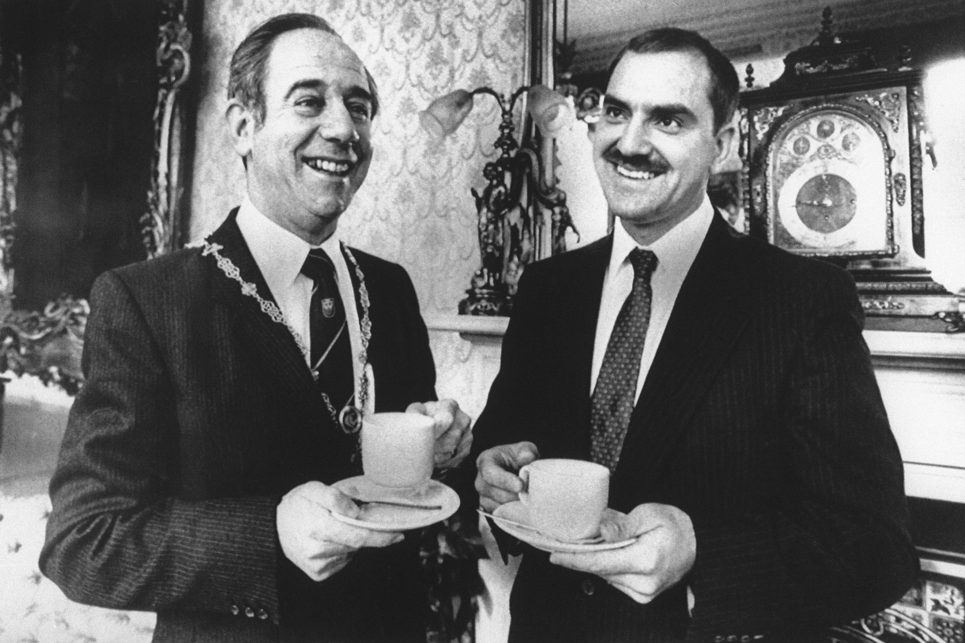 two men in suits smile while holding teacups in an ornately-decorated room