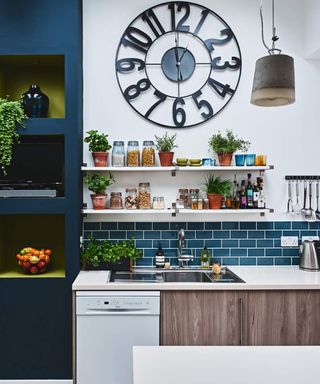 A modern blue kitchen with white dishwasher appliance, large black metal clock decor on wall and tiled blue backsplash behind metal sink