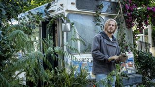 Ish (Alexander Ludwig) stands in front of an abandoned bus overgrown with plants in Earth Abides