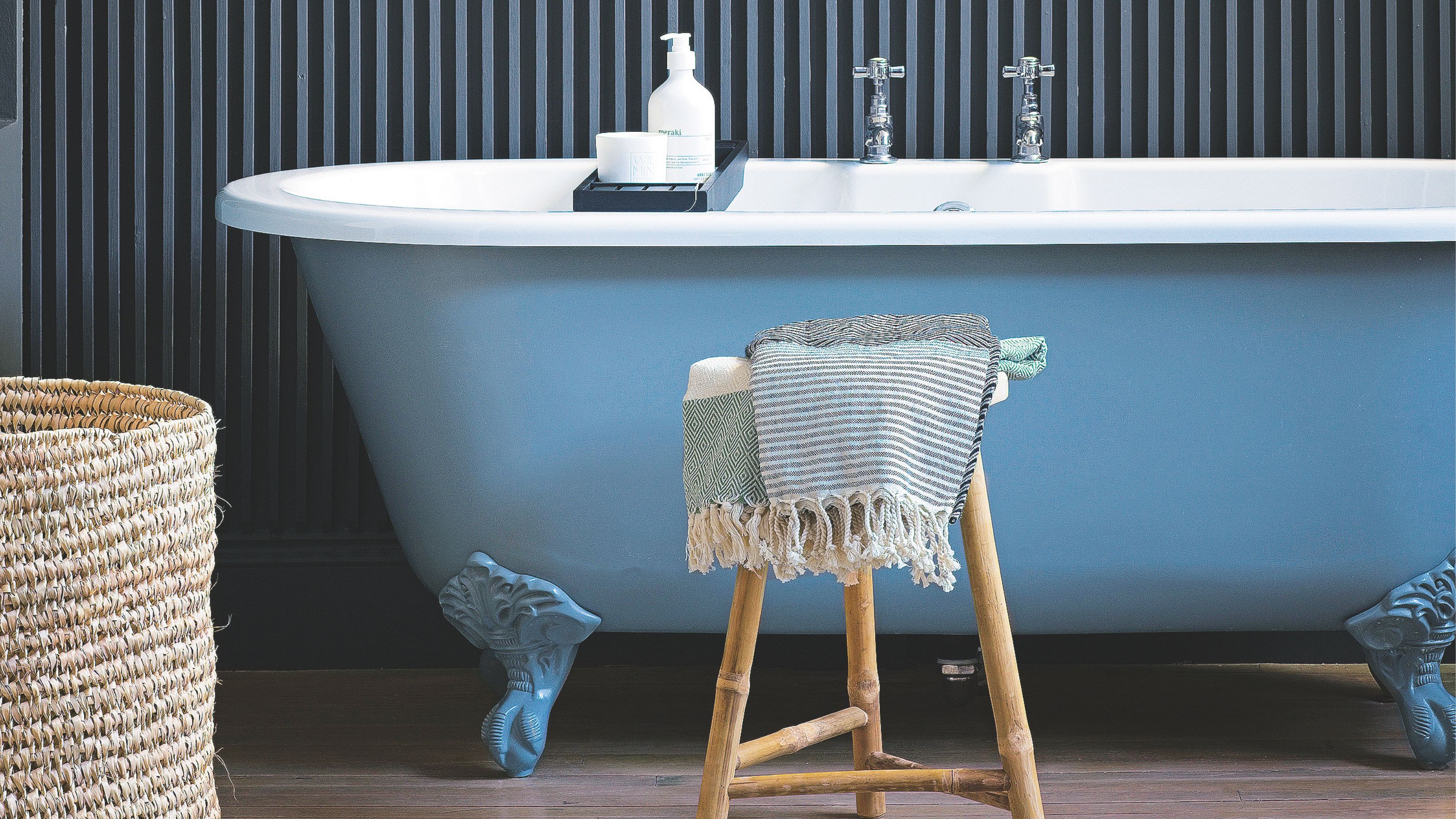 Blue and white freestanding bathtub, in front of dark blue clad wall, and a wooden stool with tassled blanket on it