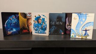4K Blu-ray cases of Tron, Rocky Horror Picture Show, Fantastic Four: First Steps, Se7en, The Warriors and The Sound of Music in a line on dark wood table 