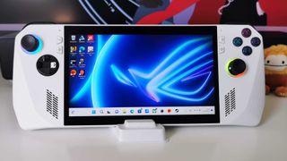 Asus ROG Ally sitting on white desk with stand and Squishmallow big foot on right hand side.