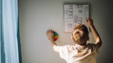 A boy with blond hair reaches up to fix a weekly calendar.