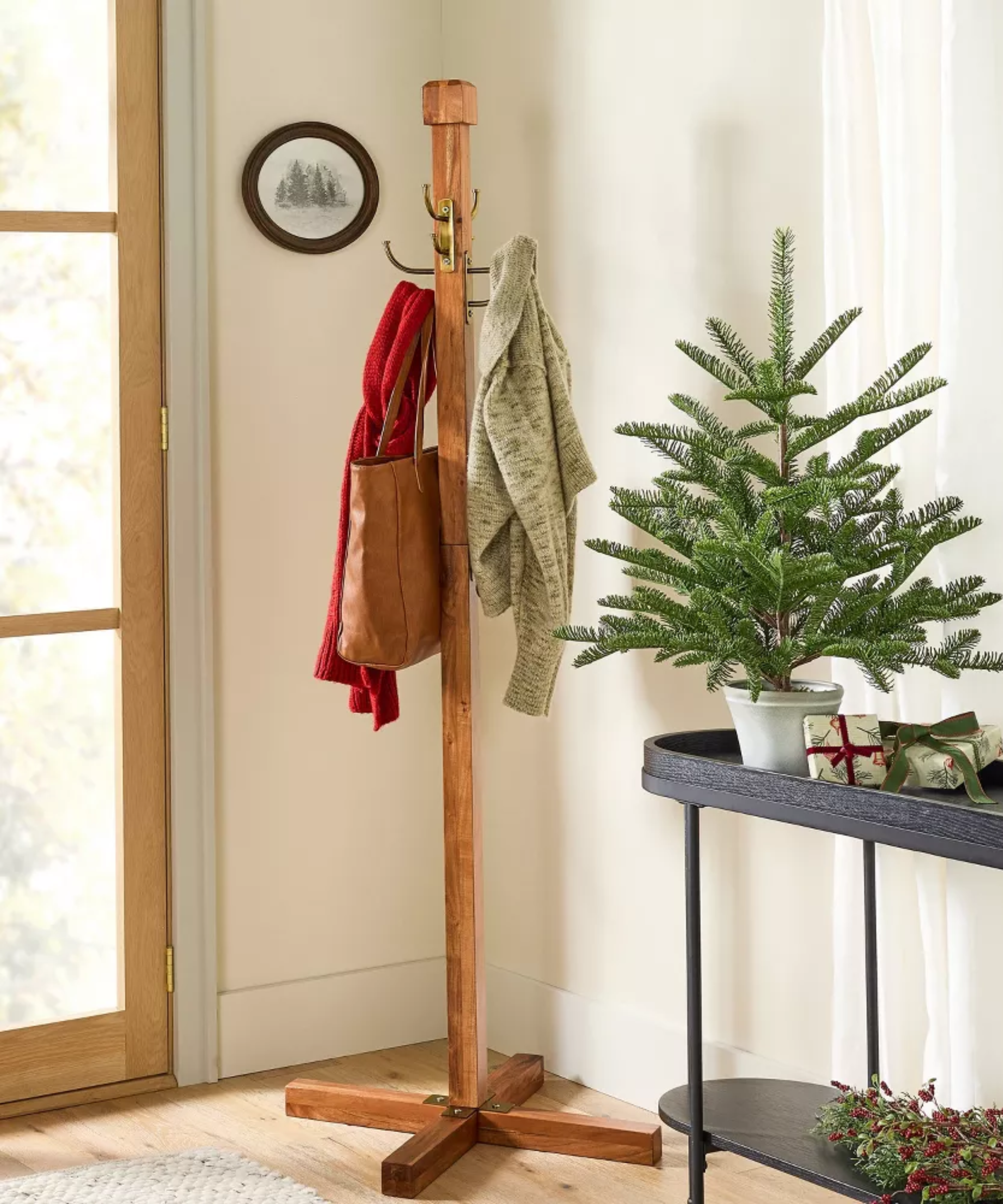an entryway with a wooden coat rack holding two coats and a bag, and a small black table holding a small tree and two wrapped gifts