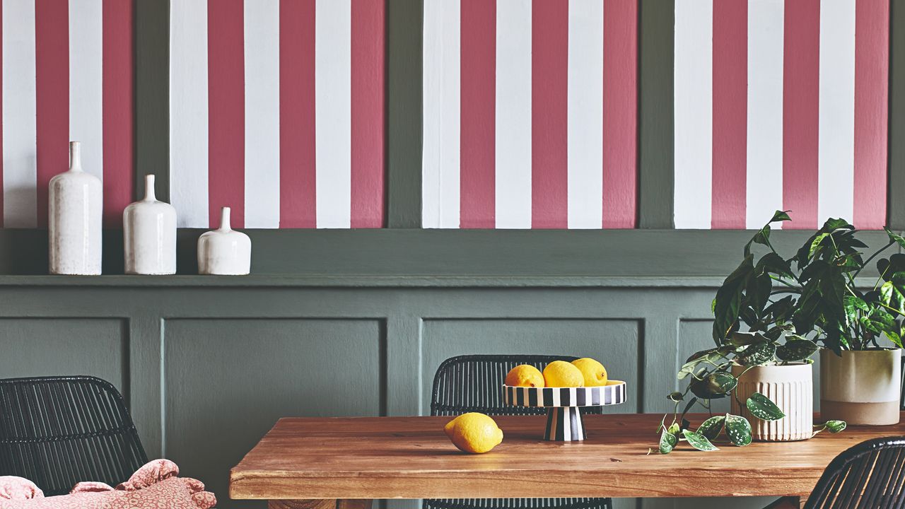 A dining room with a wall covered in a red and white striped wallpaper and green panelling with a striped fruit bowl on the dining table