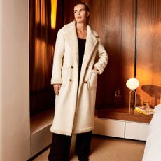 Model infront of minimal wooden interior wearing a white long collared afghan coat sold at Warehouse 