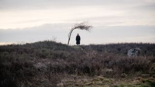 An old woman in the distance under a sparse windswept tree.