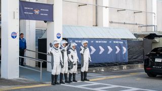 astronauts walk out of a building and board black cars.