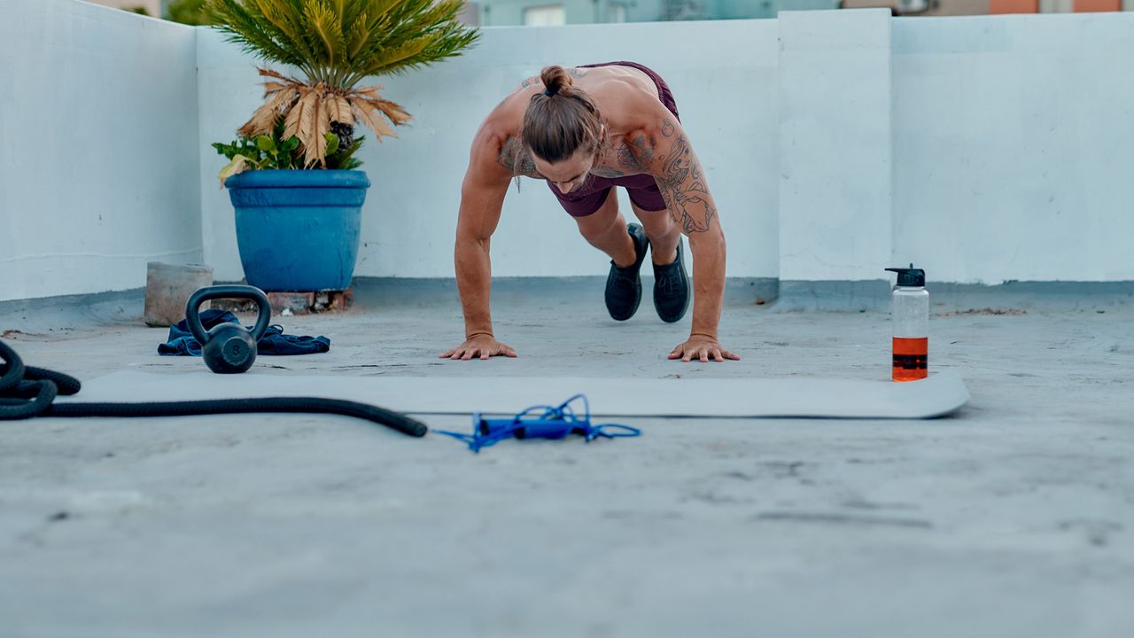 Man performing a high plank burpee during an outdoor workout with exercise mat and equipment around him
