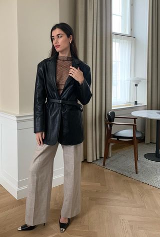 A woman wearing a black leather blazer over a sheer textured top styled with tan linen trousers and black slingback heels.