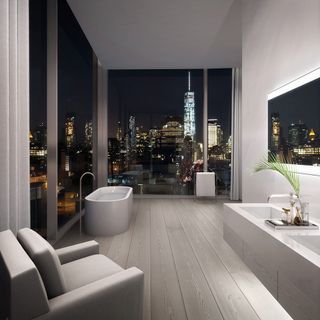 A bathroom in an apartment featuring white oblong shaped bath against glass walls with a mirror on the opposite side of the wall and a grey sofa in a corner of the bathroom