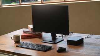 An MSI Cubi NUC 1M mini Pc on a wooden desk next to a monitor with a blank screen. A wireless laptop and mouse are in front of the monitor and mini PC, while a white mug with a brown bottom is to the left of the keyboard. Three books – The Circle, Nordic Interior Homes, and another the title of which is obscured – are arranged to the left and slightly behind the monitor. The PC and all peripherals are black,