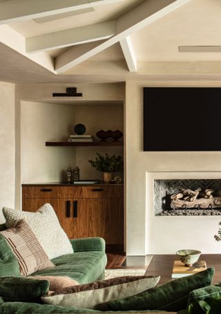 Living room with beige plaster-effect walls and ceiling, light painted ceiling beams, green velvet sofa and open fireplace