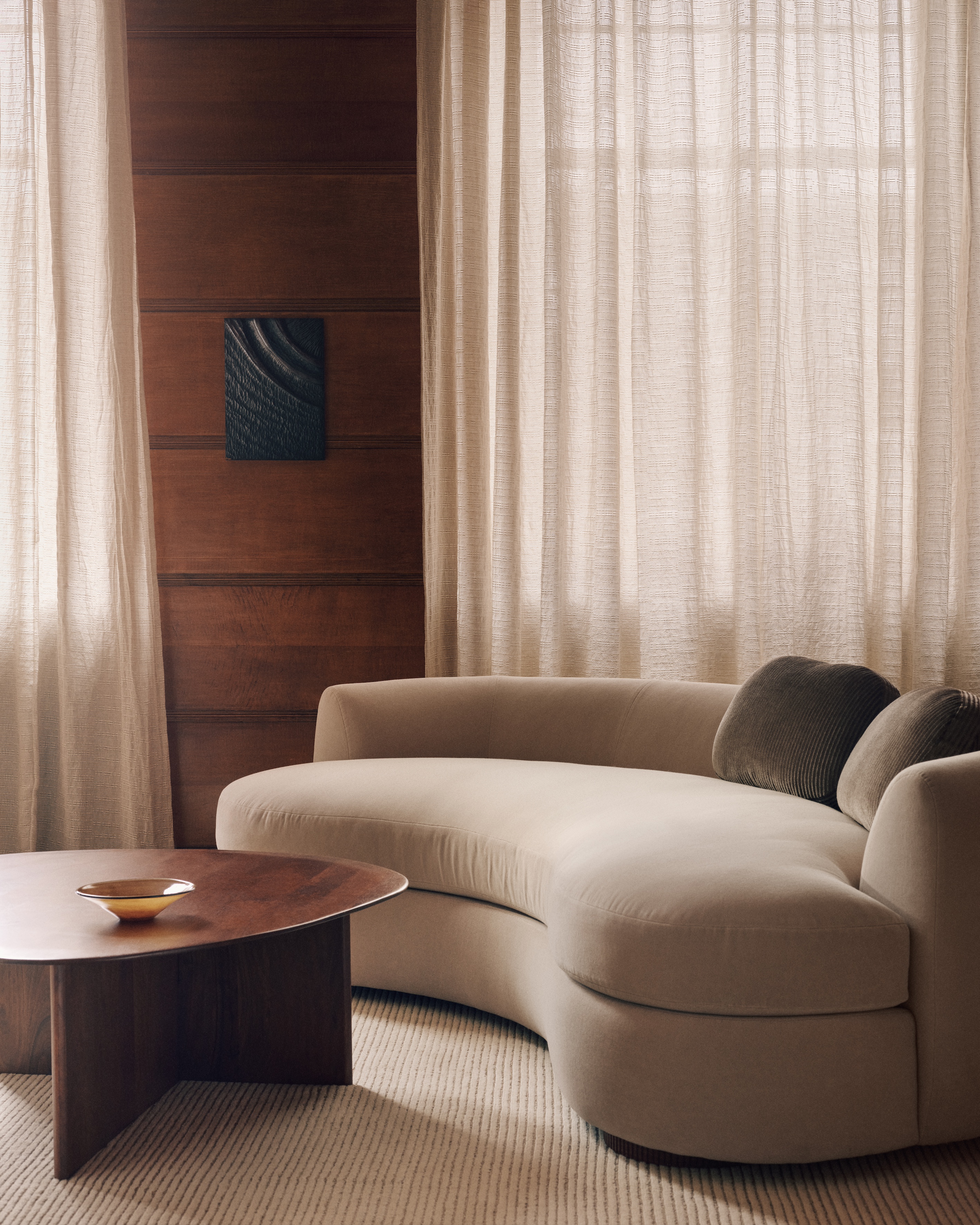 A wood-panelled living room with two windows that are covered in sheer linen curtains. There is a curved off-white sofa with an off-white rug underneath and a round wooden coffee table.