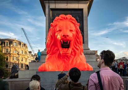 View of Please Feed The Lions by Es Devlin at Trafalgar Square during the day - a red lion statue with a black display in its mouth that is showing poetry. There are people standing around the lion and buildings in the background