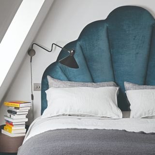 A bedroom with a bed with a large curved velvet headboard and a stack of books on the bedside table