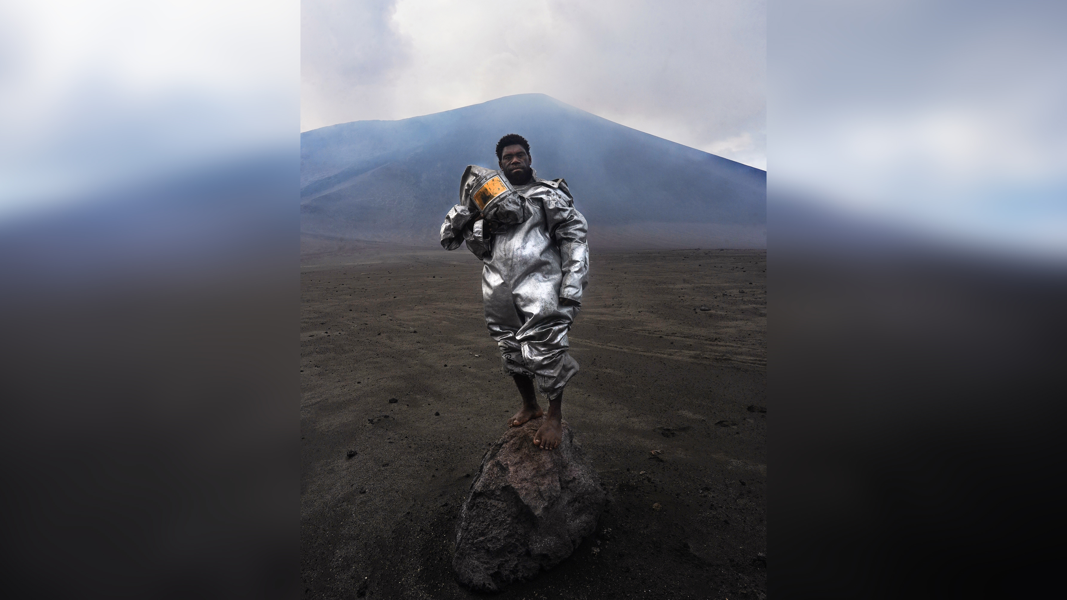 Phillip, a self-taught volcanologist, stands barefoot atop a volcanic rock bomb. Wearing a lava-protection suit, the volcano smoulders behind him, sending a plume of gas and sulphur into the sky
