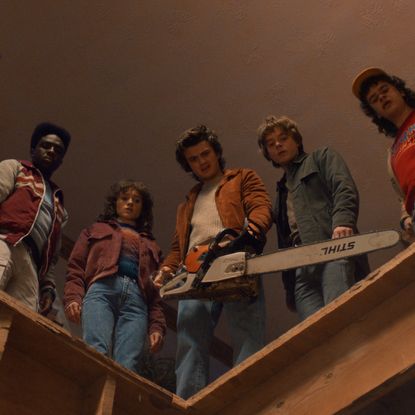 A group of teenagers (l-r: Finn Wolfhard as Mike Wheeler, Caleb McLaughlin as Lucas Sinclair, Natalia Dyer as Nancy Wheeler, Joe Keery as Steve Harrington, Charlie Heaton as Jonathan Byers, and Gaten Matarazzo as Dustin Henderson) look down from a wooden ledge, as one man holds a chainsaw, in STRANGER THINGS: SEASON 5.