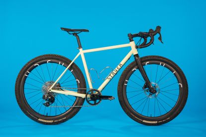 The Stayer Groadinger UG gravel bike is pictured here side on. The handle bars point to the right, it is a grey/green colour frame and is shot on a blue background