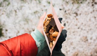 View from above of an open bag of nuts and squares of chocolate. It is held underneath by a person's hand. The arm of a red jacket comes in from the bottom left of the image.