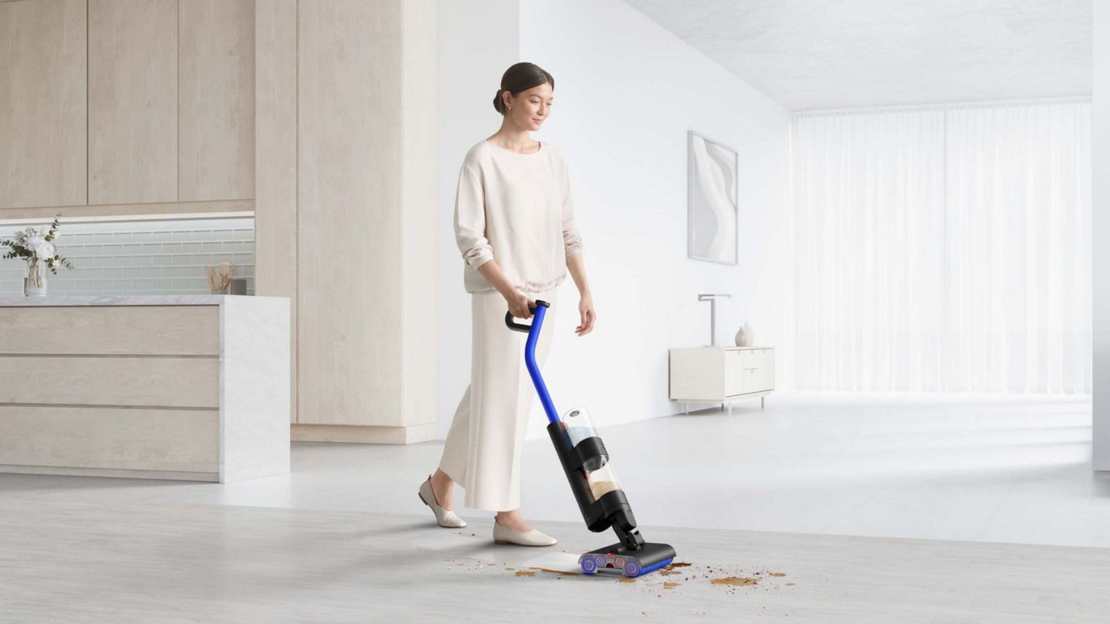 A women with brown hair in a low bun wearing a white long-sleeved top, trousers and shoes, using the Dyson WashG1 to clean up a spillage in a large modern white home space, with kitchen cabinetry to the left, and a large empty living room to the white. In the background are large windows with white curtains.