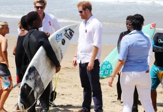 Prince Harry wearing a white shirt and black pants standing on a beach talking to someone holding a surfboard