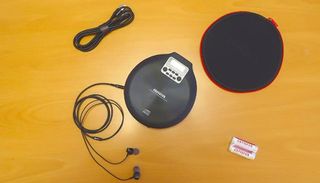 A top-down shot of the Aiwa PCD-810BL portable CD player with attached in-ear headphones, carry case, cable and 2 small batteries on a mustard-yellow tabletop.