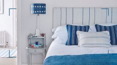 White bedroom with blue coastal-style with a linen pinstripe headboard to suggest how to keep a bedroom cool 