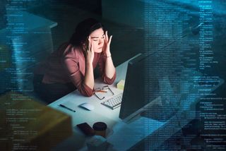 A shot of a woman sat at her desk in a dimly lit office, with her eyes closed and a stressed expression on her face, her hands raised to massage her temples. In the foreground, blue code is rising to either side of the frame to indicate complexity in the task that her unseen screen is showing