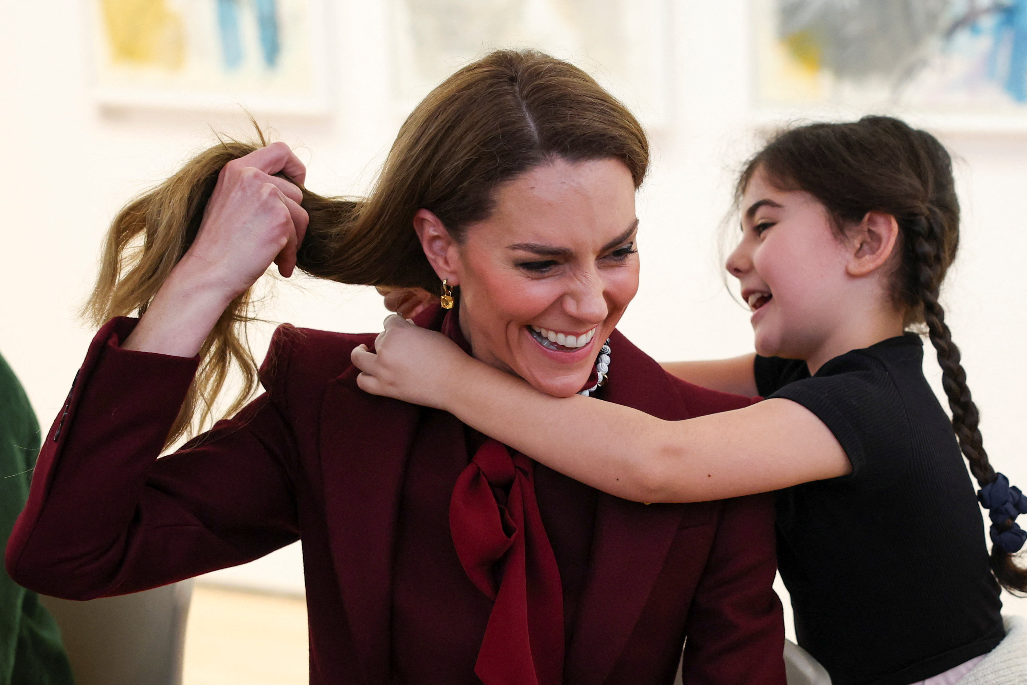 Princess Kate wearing a burgundy outfit, lifting her hair for a little girl to tie a necklace around her neck.