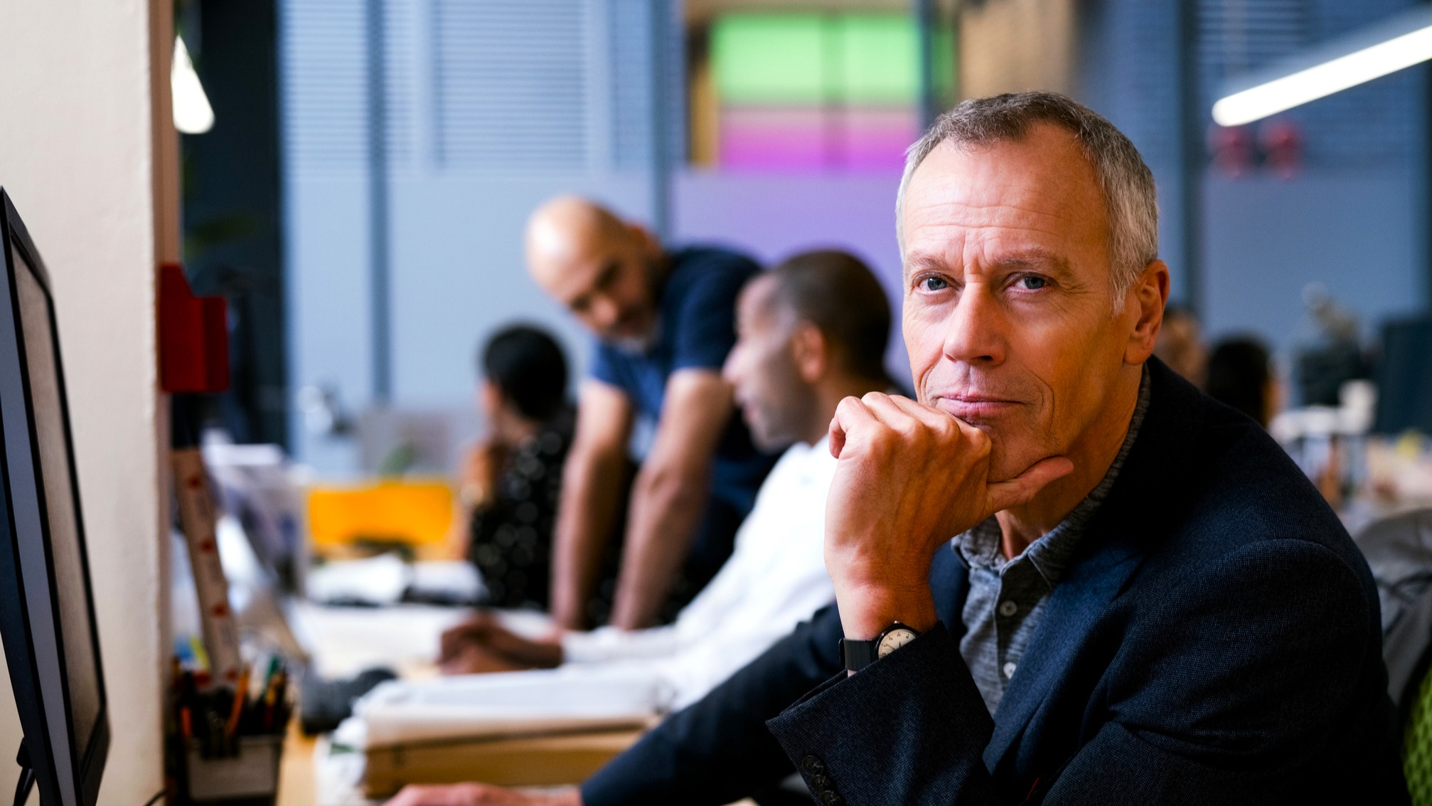 Portrait of senior businessman with hand on chin at desk. Confident male professional is using computer. He is with colleagues in creative office.