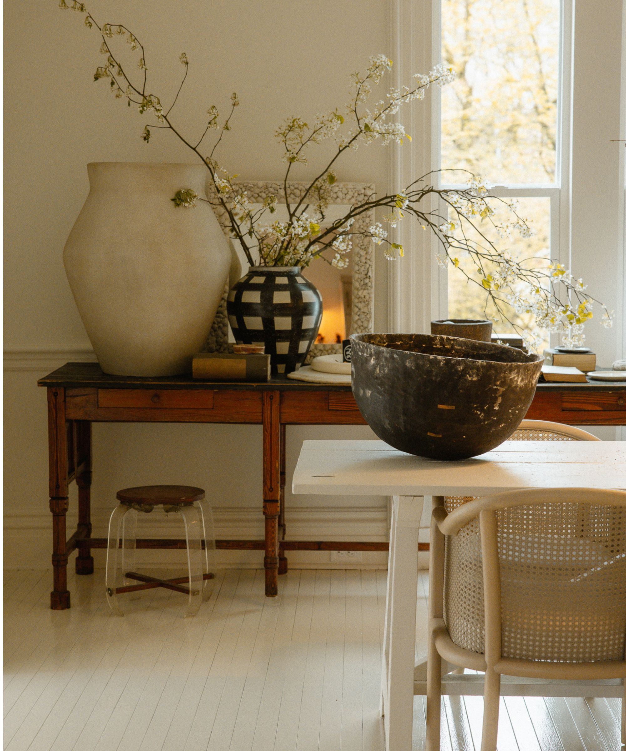 Dining room with white wooden floor, large window, wooden console underneath the window with large vases and branches, and a white dining table with a rustic bowl and rattan chairs underneath