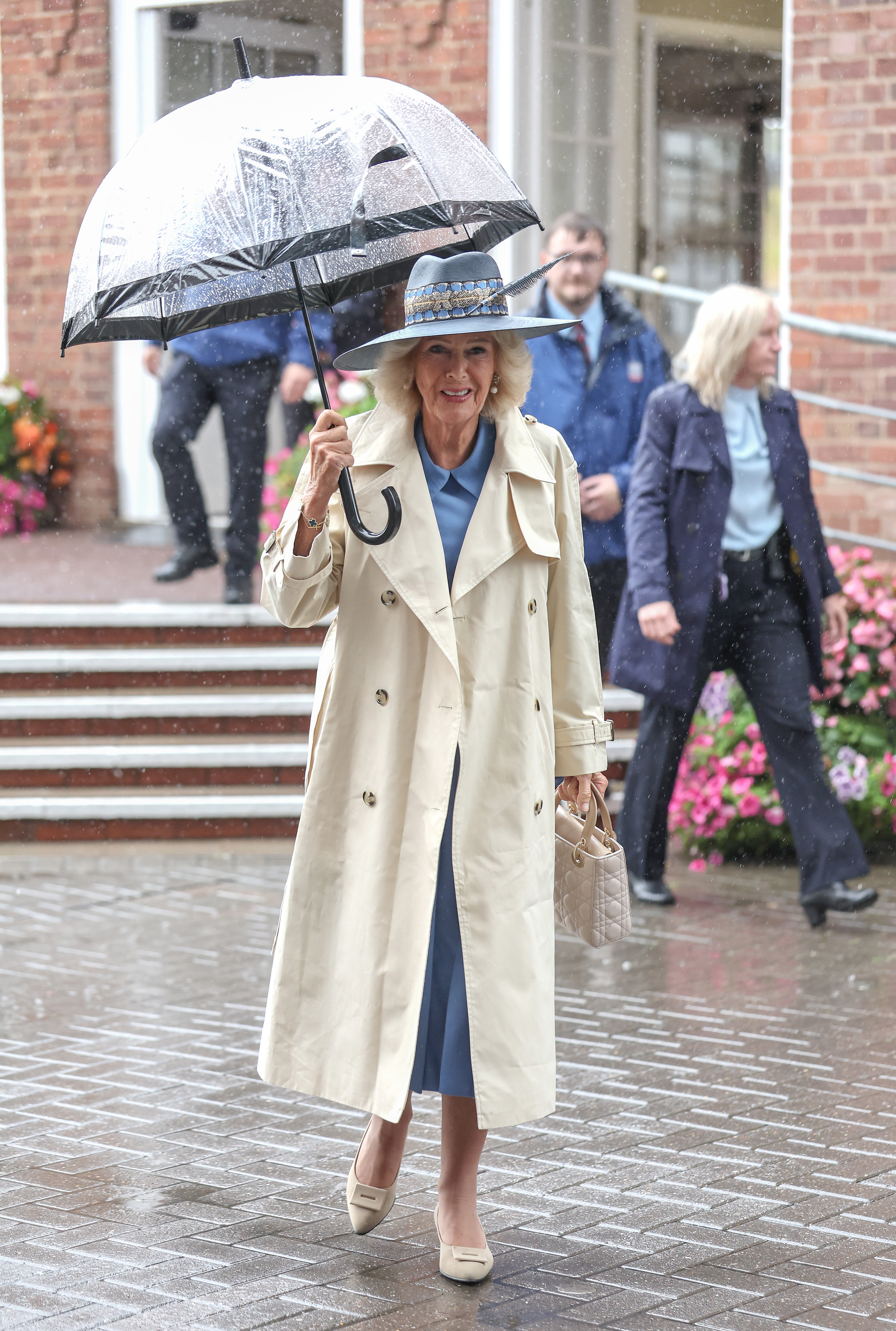 Queen Camilla walking in the rain wearing a trench coat and holding an umbrella