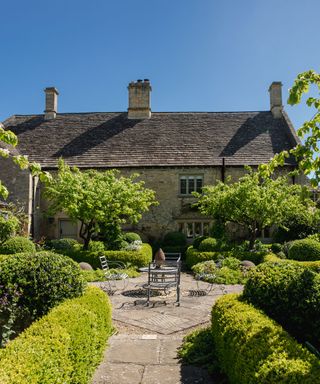 A formal courtyard garden with stone pavers, box hedges, white flowers and an iron table and chair set in front of a stone country cottage.