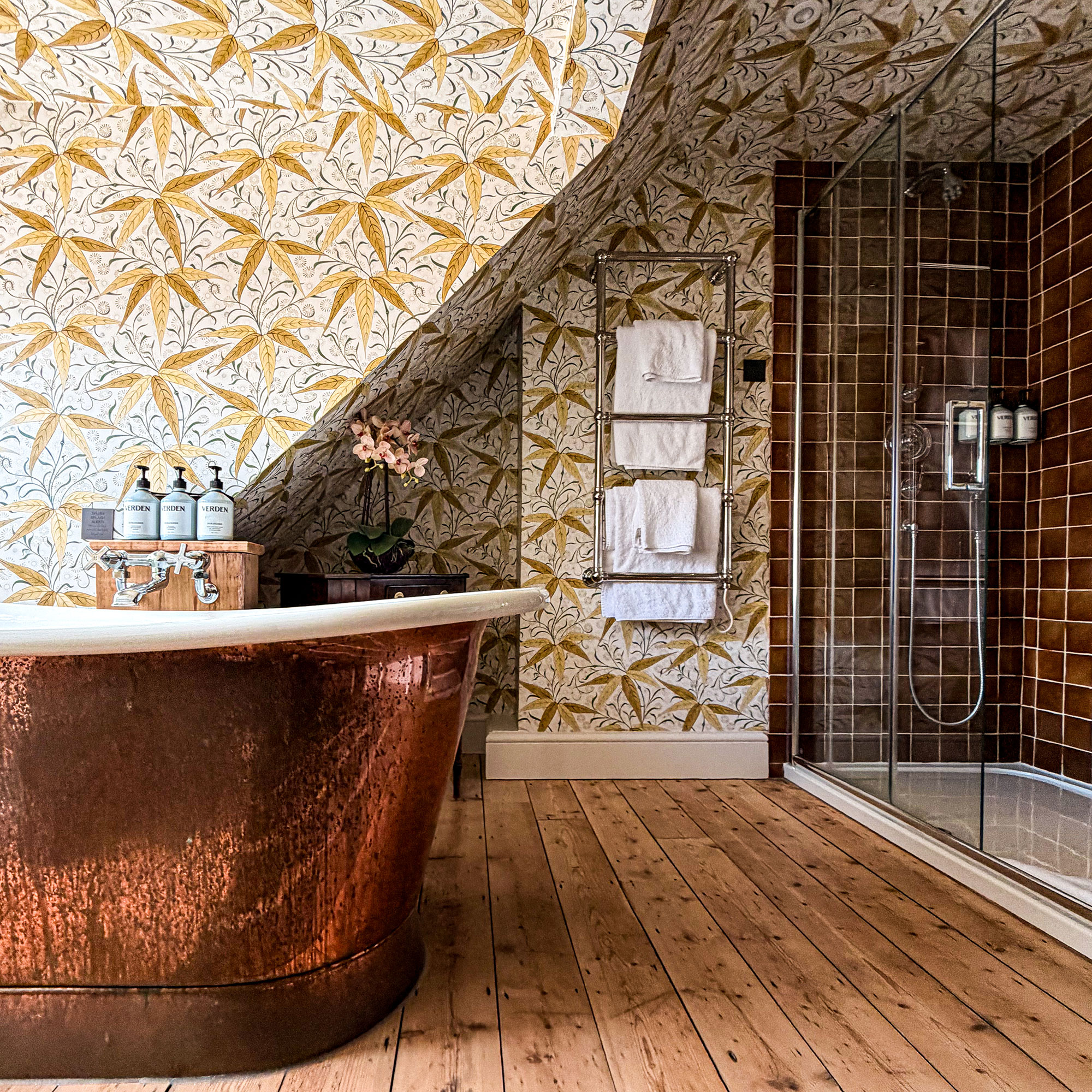 Bathroom in eaves covered in patterned yellow wallpaper, with copper roll top bath, wooden floor and walk-in shower with brown tiles