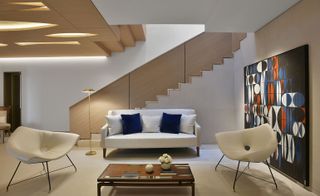 Communal area with cream sofa and two chairs. Coffee table in the middle in front of a piece of geometric artwork and a staircase in the background
