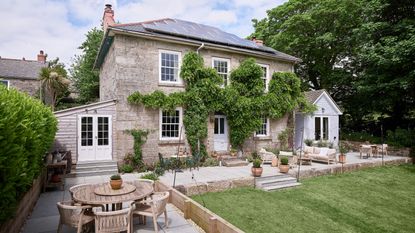 Exterior shot of the back of a Georgian farmhouse with wisteria growing up walls, solar roof panels, patio and lawn area with outdoor furniture