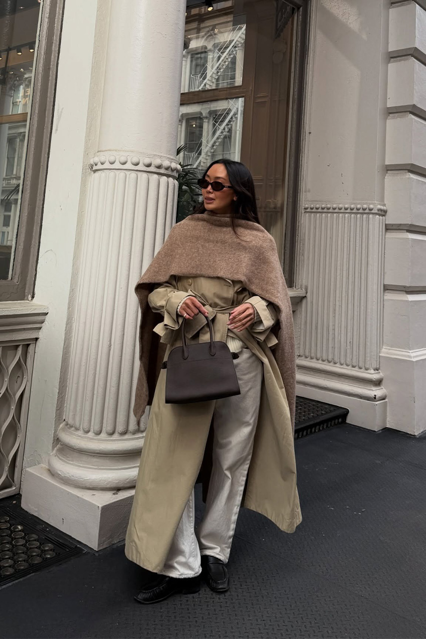 West Village winter trends are shown in a street-style photo of a woman standing in front of a white, glass storefront in her winter outfit. She's wearing a tan trench coat with a long brown scarf wrapped around the shoulders like a cape, styled with a brown bowler bag, white baggy jeans, brown loafers, and brown sunglasses
