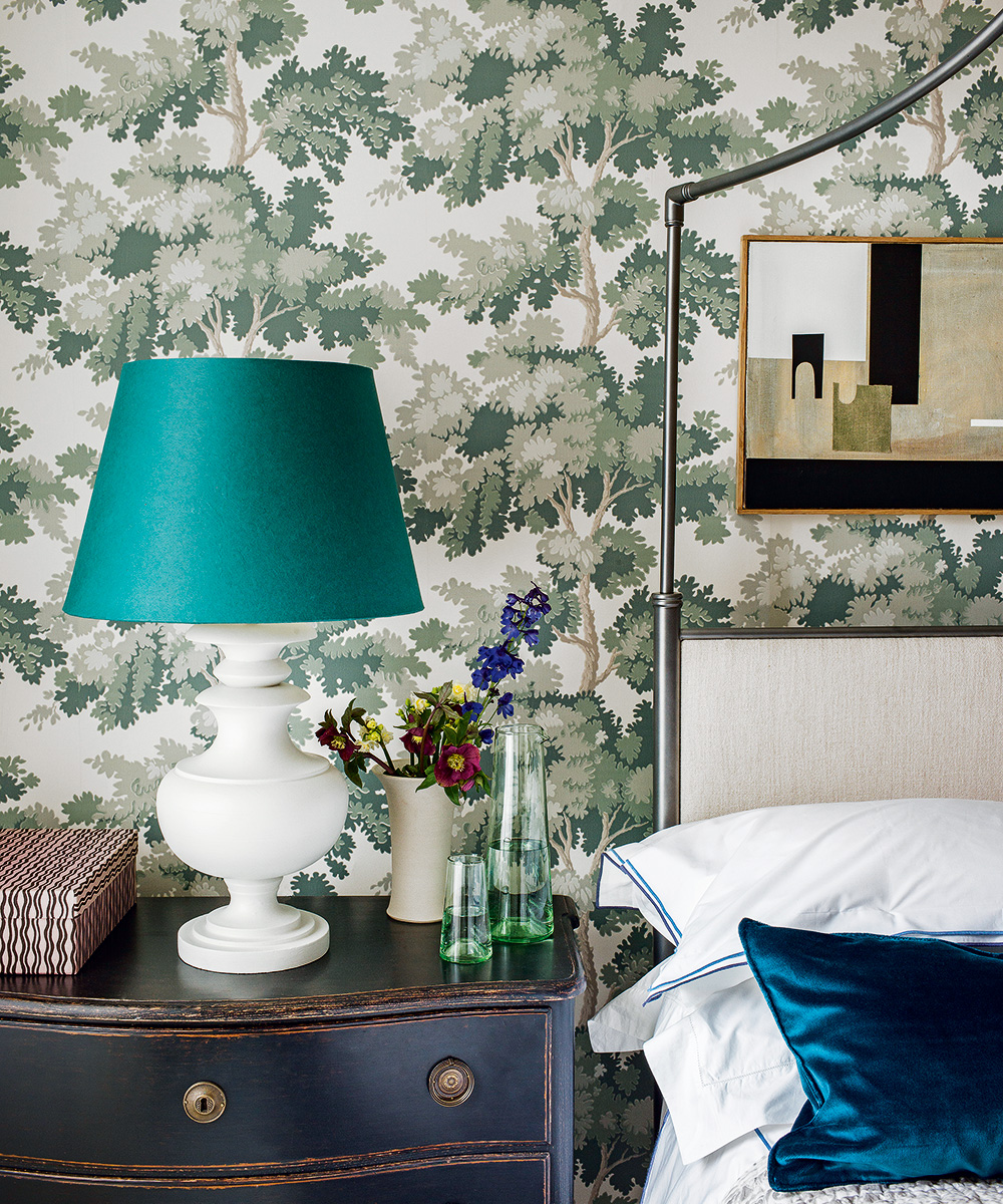 Green and white tree-print wallpaper in a traditional bedroom with antique drawers and a white and blue lamp beside a metal bed.