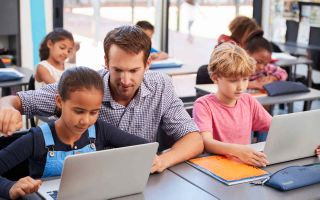 Male teacher works with a boy and a girl at laptop computers in the classroom.