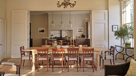 a large warm open kitchen and living room in a Georgian home with dark green cabinetry and AGA cooker seen through kitchen doors, an antique wood table and chairs sit in front 