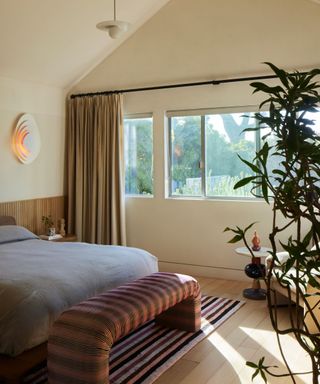 Neutral bedroom with large windows and dark beige drapes, bed with gray bedding and rattan bench