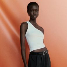 Woman wearing white one-shoulder tank top, and black flowy skirt with leather belt against orange studio backdrop.