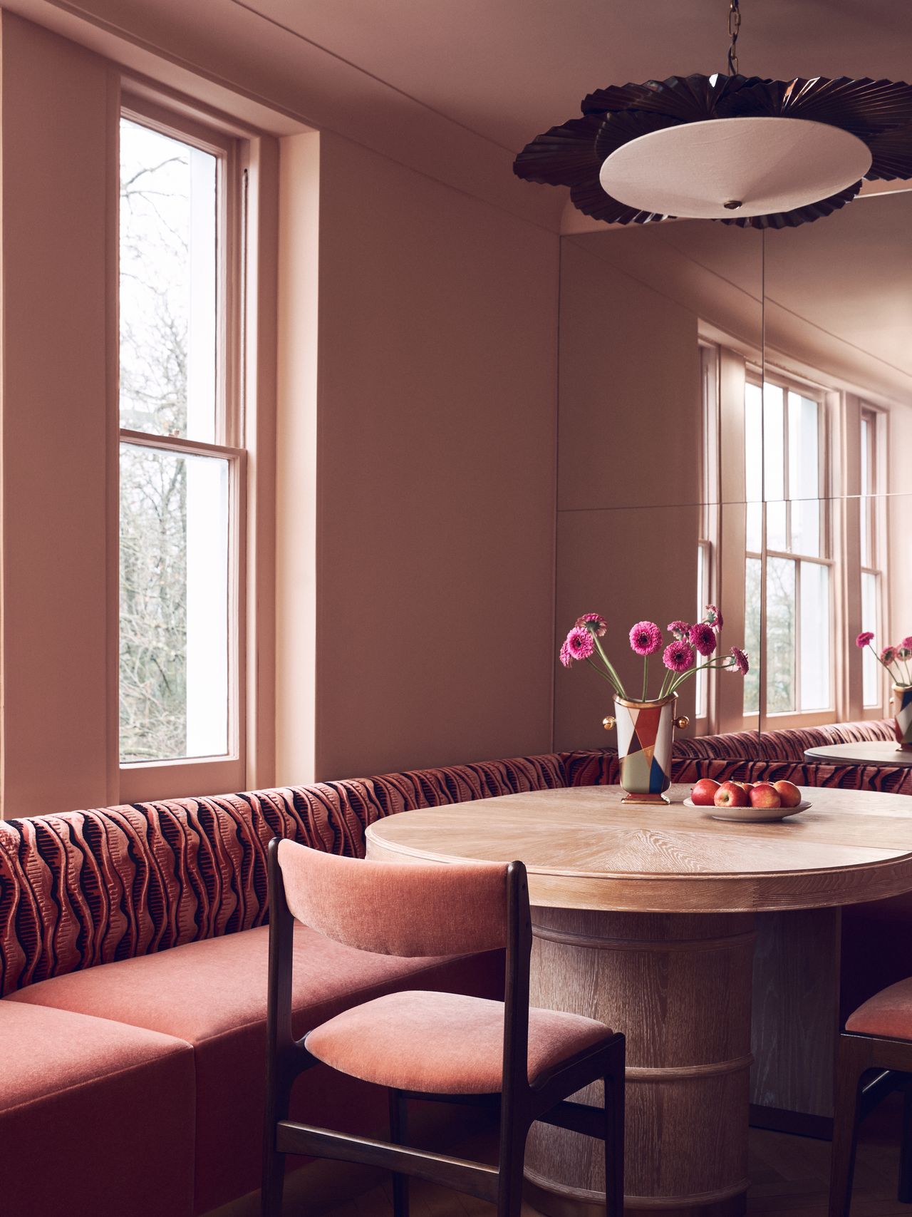 Image of a dining nook drenched in light, dusty pink. There is a pink bench seat with a patterned back, a round wooden table, a mirrored wall, and matching pink dining chairs.