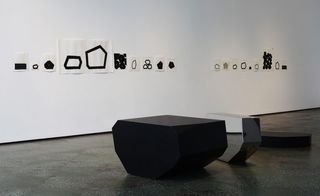 White papers with black geometrical shapes drawn on them are displayed on the wall. There is one black and one reflective asymmetrical structure on the floor.